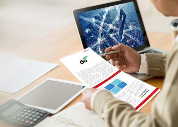 Person at a desk holding a document about 5G, with a laptop displaying an image of a connected city in the background, alongside a tablet and a calculator.